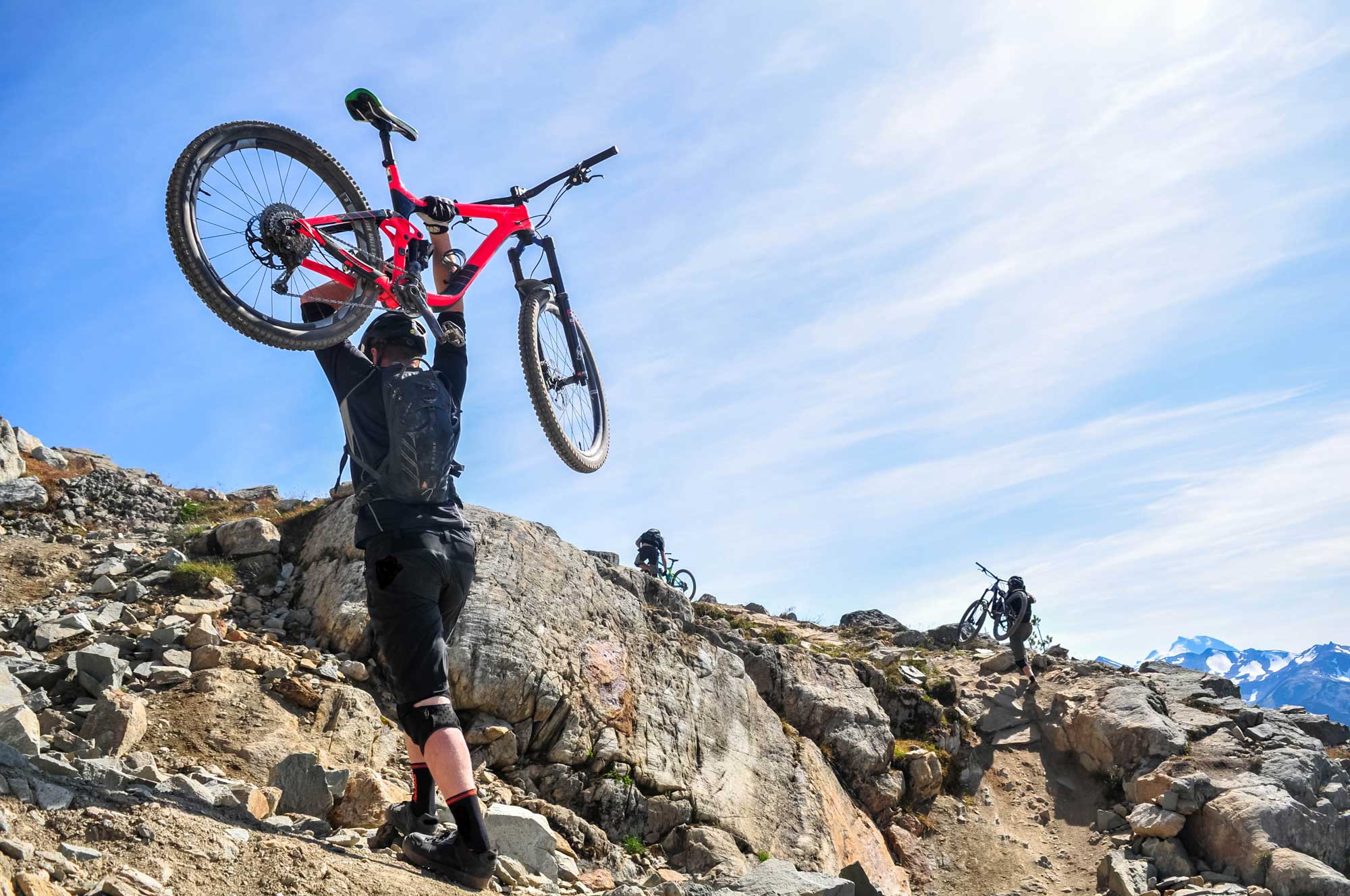 on rocky terrain, a man holds a red mountain bike over his head; two more mountain bikers ride in the background